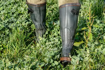 Eine Person geht in schwarzen Stiefeln durch das hohe Gras einer feuchten Wiese.